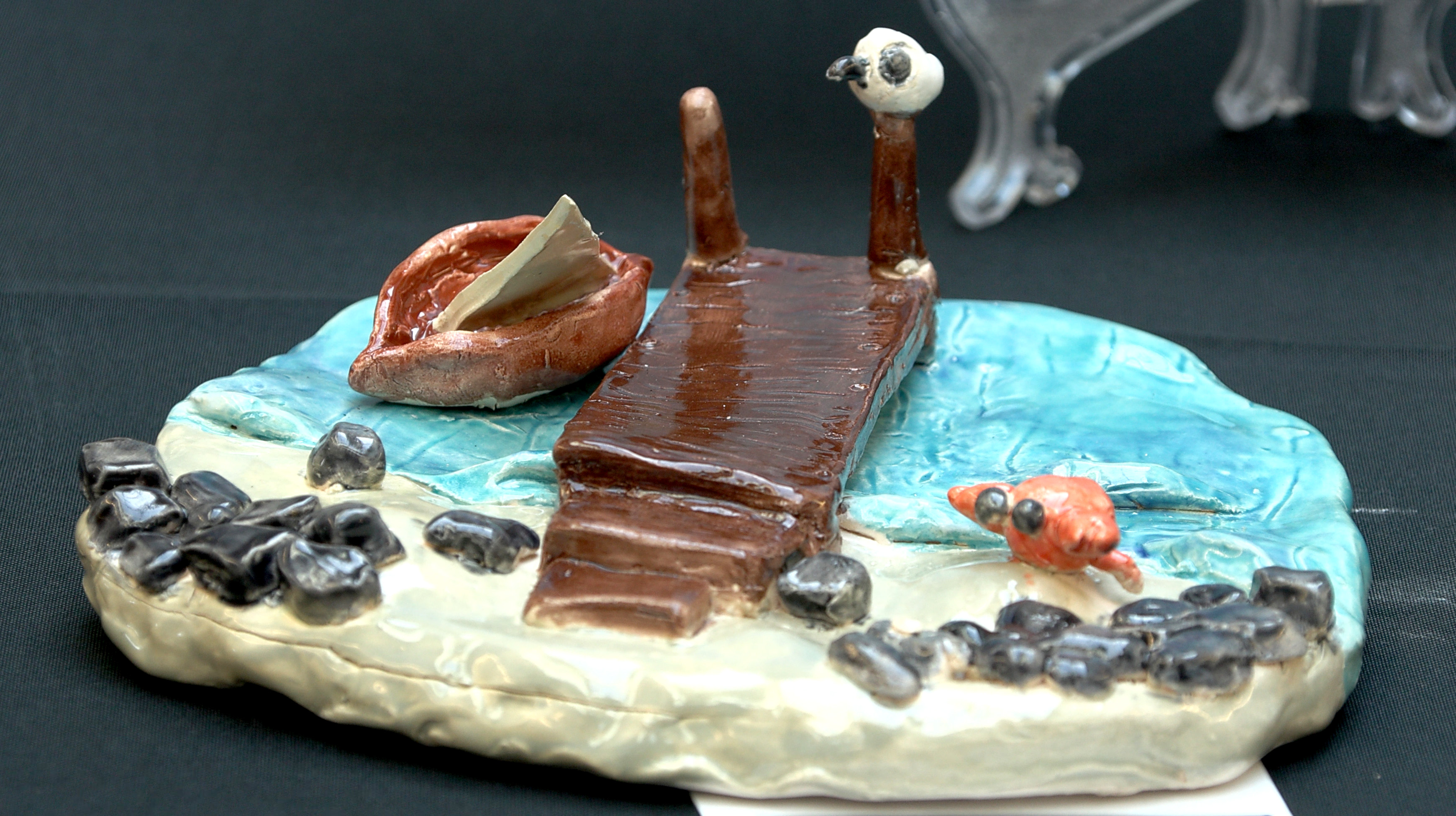 Student created ceramic art of a dock and canoe on a rocky beach with a crab and seagull.